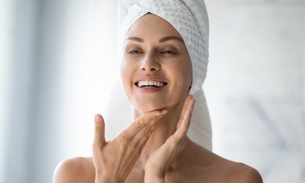 Happy woman with her hair wrapped in a towel looking at her liquid facelift results in the bathroom mirror.