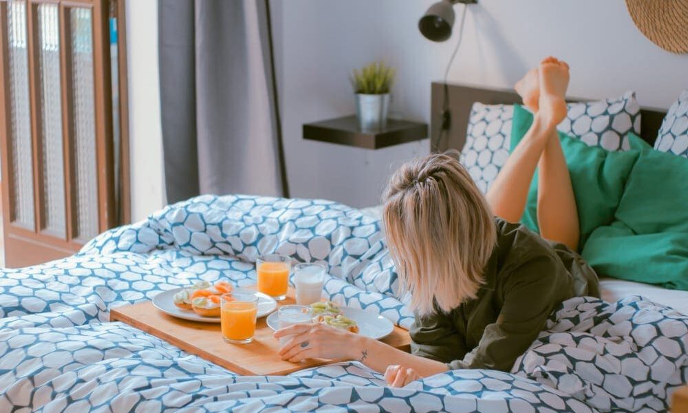 Woman relaxing on her bed while eating from a food tray.
